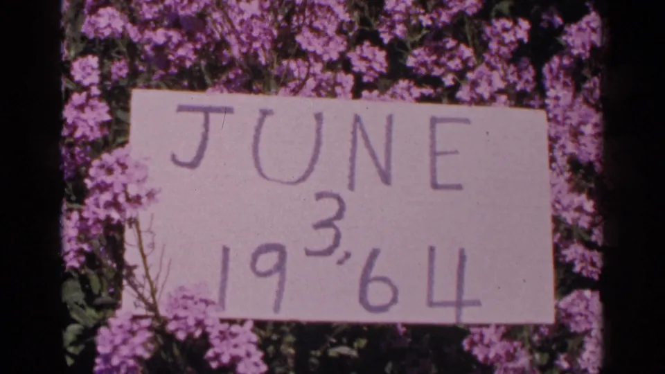 A close view of a handmade June 3, 1964 sign set against flowering shrubs in the dated home-movie sequence.