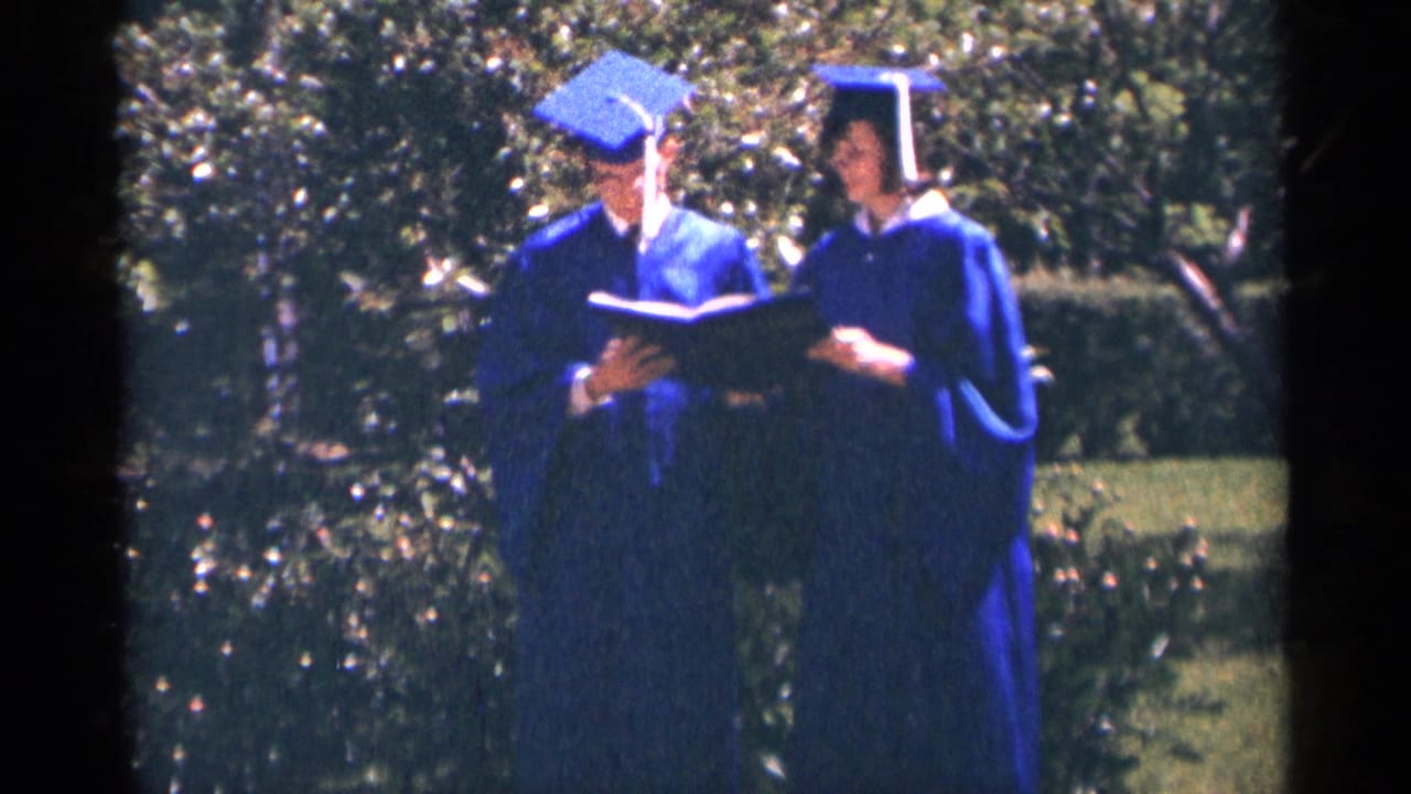 Two graduates in blue caps and gowns stand outdoors in Minnesota, holding papers amid greenery in 1964 home-movie footage.