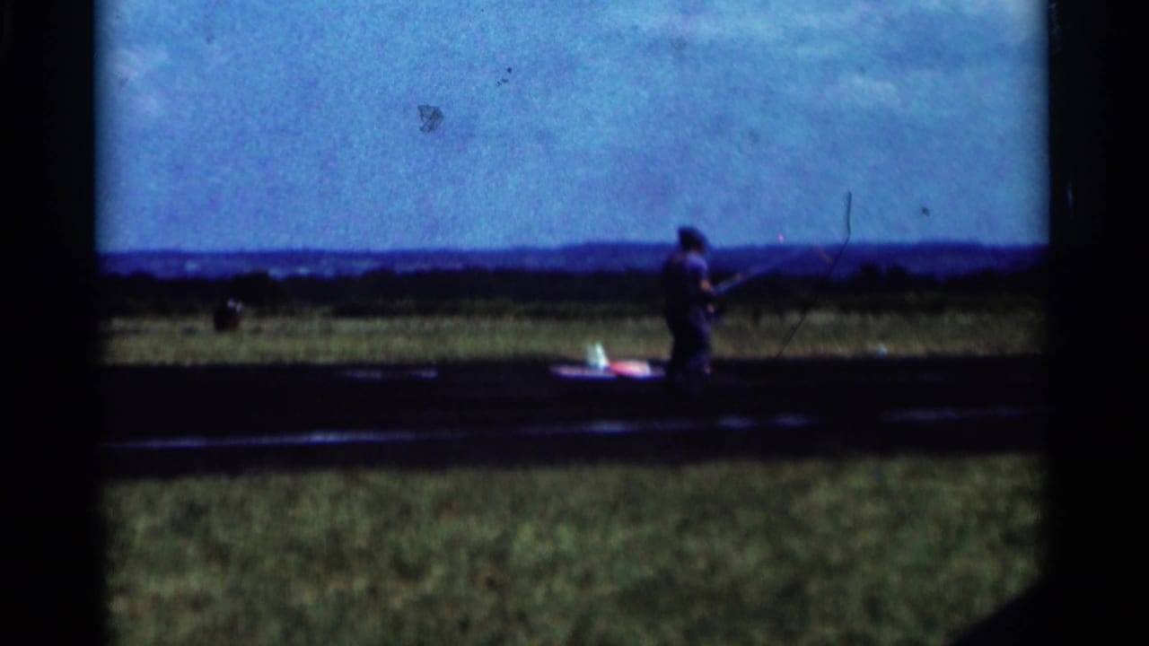 Man near a small model airplane on a grassy airfield under a blue sky.