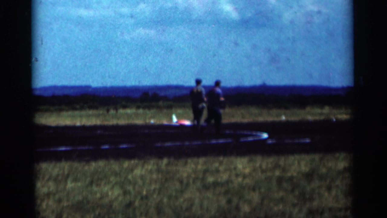 Two men walk near a small model plane on the edge of an airfield.