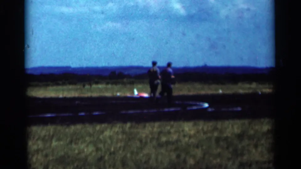 Two adult men walk across a Minnesota airfield near a red and white model airplane in this 1964 home-movie frame.