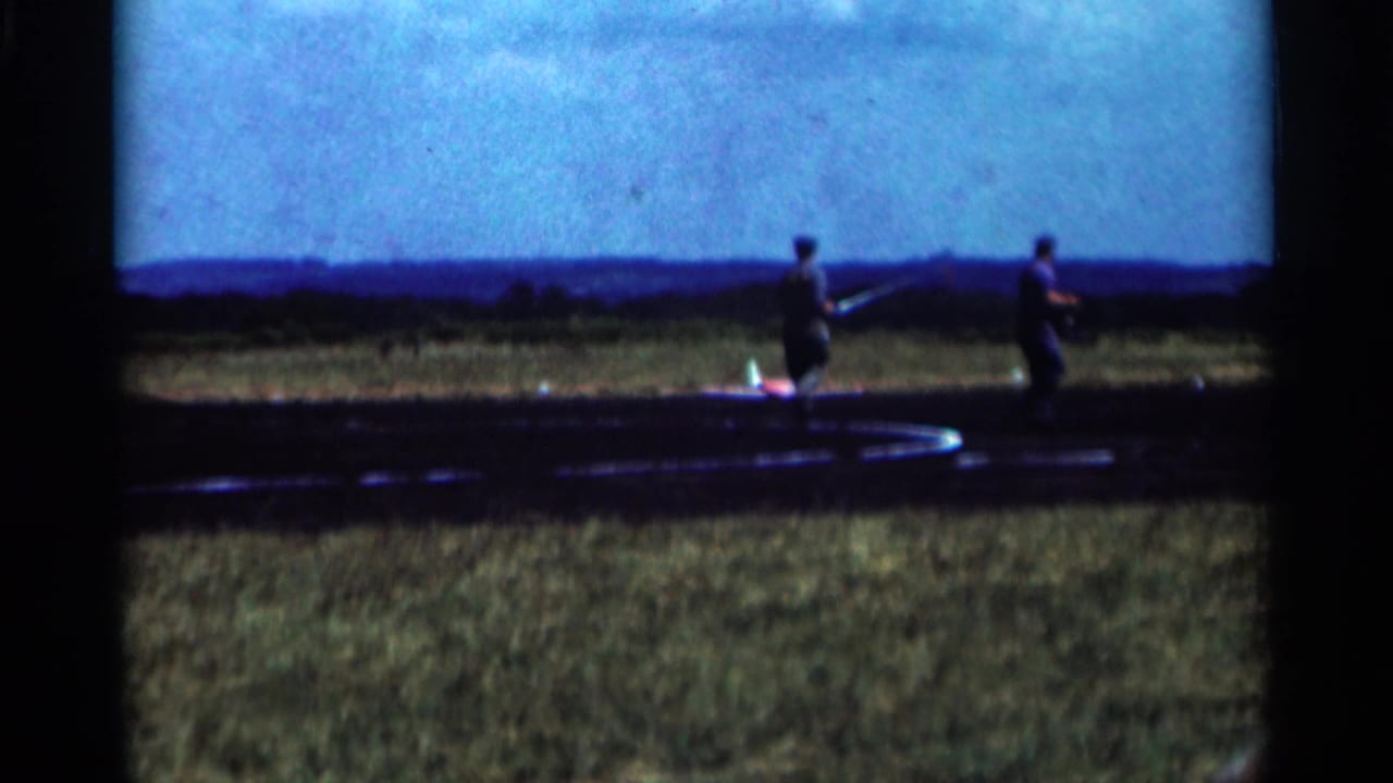 Two adult men are seen near a red and white model aircraft on a 1964 Minnesota airfield, filmed in soft 8mm color.
