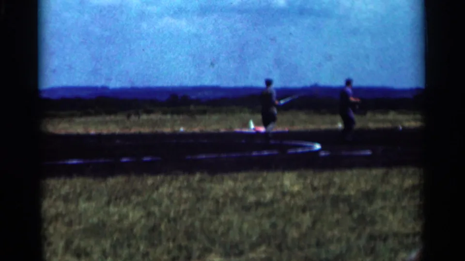 Two adult men are seen near a red and white model aircraft on a 1964 Minnesota airfield, filmed in soft 8mm color.