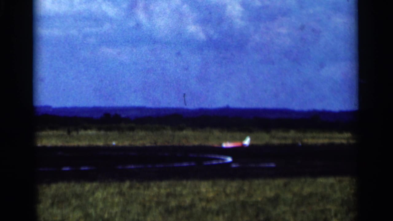 1964 Minnesota 8mm frame showing a small propeller plane low on a runway with grassy field and distant horizon.