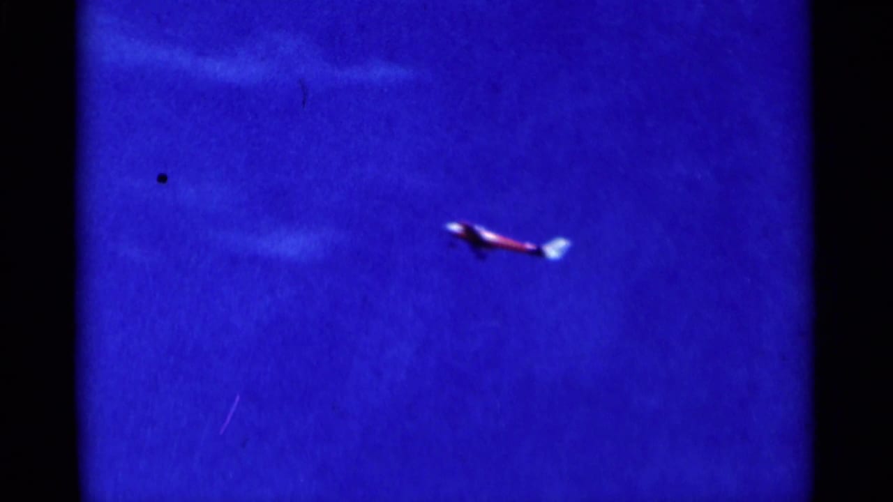 Small red model airplane crossing a deep blue sky with dark film edges.