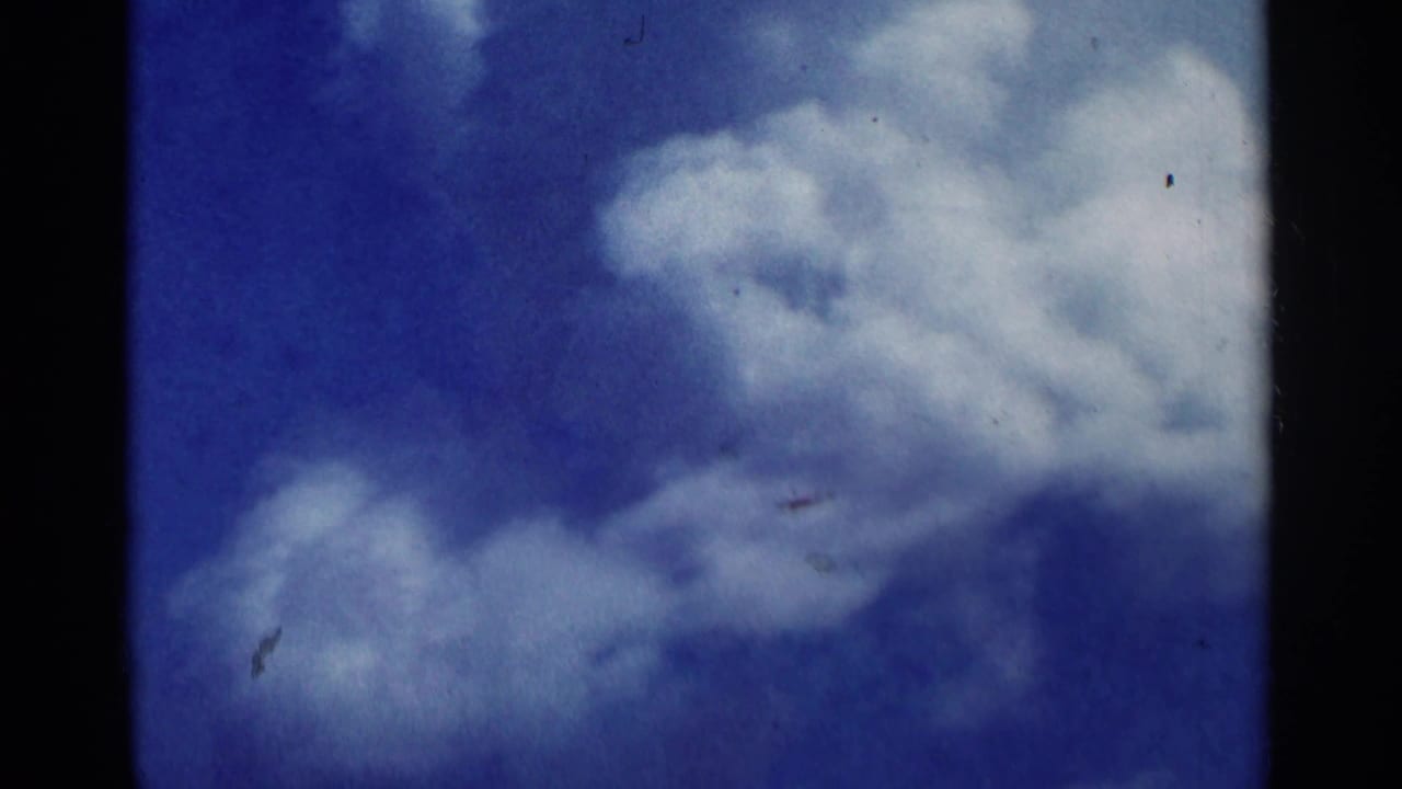 Clouds drift across a blue sky in the Minnesota home-movie clip, with film dust and dark gate edges visible.