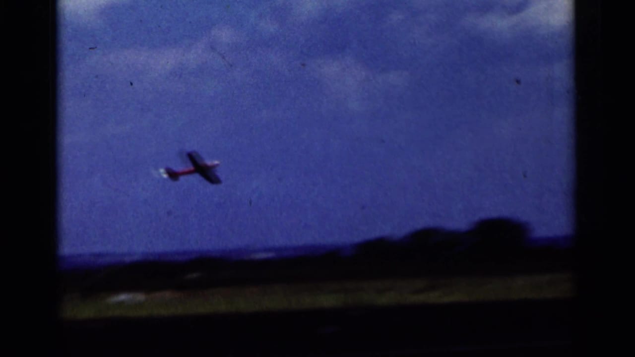 The red model airplane flies over a grassy field and distant horizon in 1964 Minnesota 8mm footage.