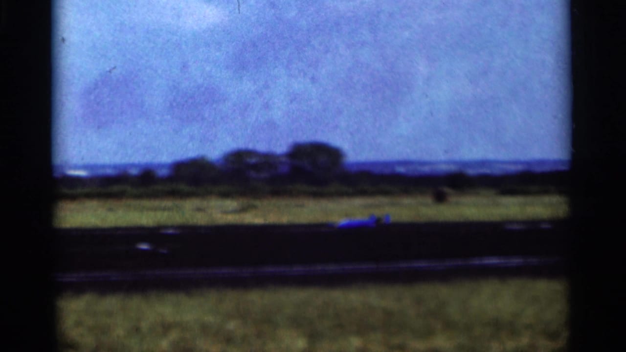 1964 Minnesota 8mm frame showing a small aircraft low on a grassy airfield with trees and horizon in the distance.
