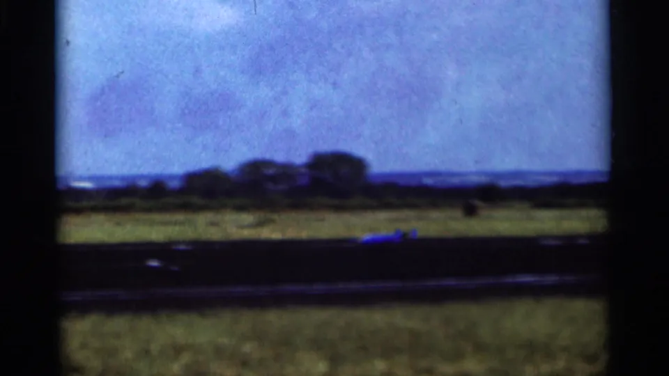 1964 Minnesota 8mm frame showing a small aircraft low on a grassy airfield with trees and horizon in the distance.