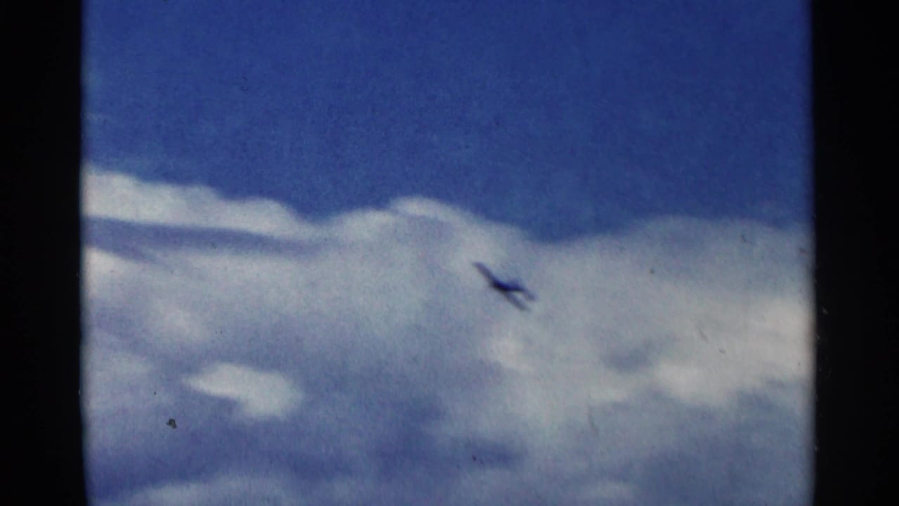 Small aircraft climbs against bright clouds and deep blue sky in 1964 Minnesota home-movie footage.