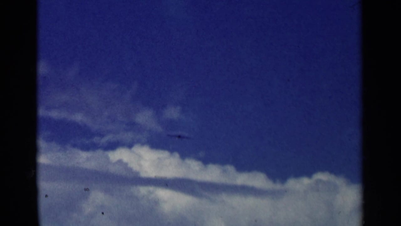 The aircraft appears as a dark shape moving across a cloudy blue sky in the 8mm aviation sequence.