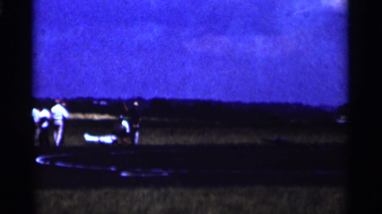 1964 Minnesota home-movie frame with onlookers near a small aircraft on a dark tarmac under blue evening light.