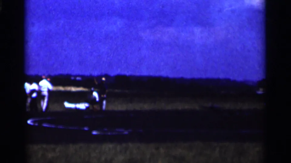 1964 Minnesota home-movie frame with onlookers near a small aircraft on a dark tarmac under blue evening light.