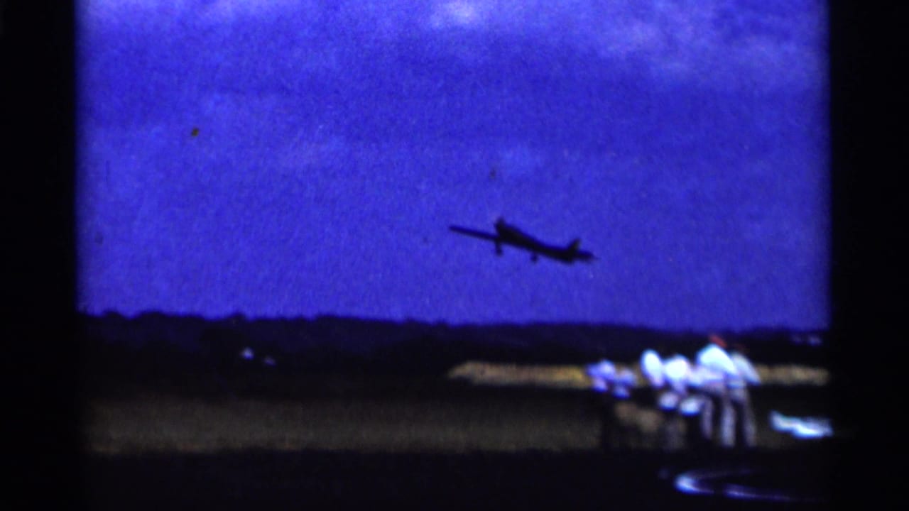 1964 Minnesota 8mm frame showing a small airplane just airborne above the airfield with onlookers below.