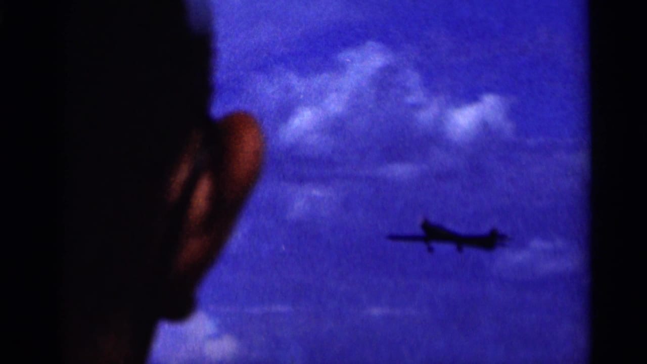 1964 Minnesota home-movie frame with a silhouetted onlooker in the foreground as a small airplane crosses a cloudy blue sky.