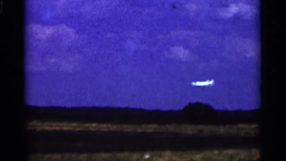 1964 Minnesota home-movie frame with a small white airplane in blue sky above a distant treeline and open field.