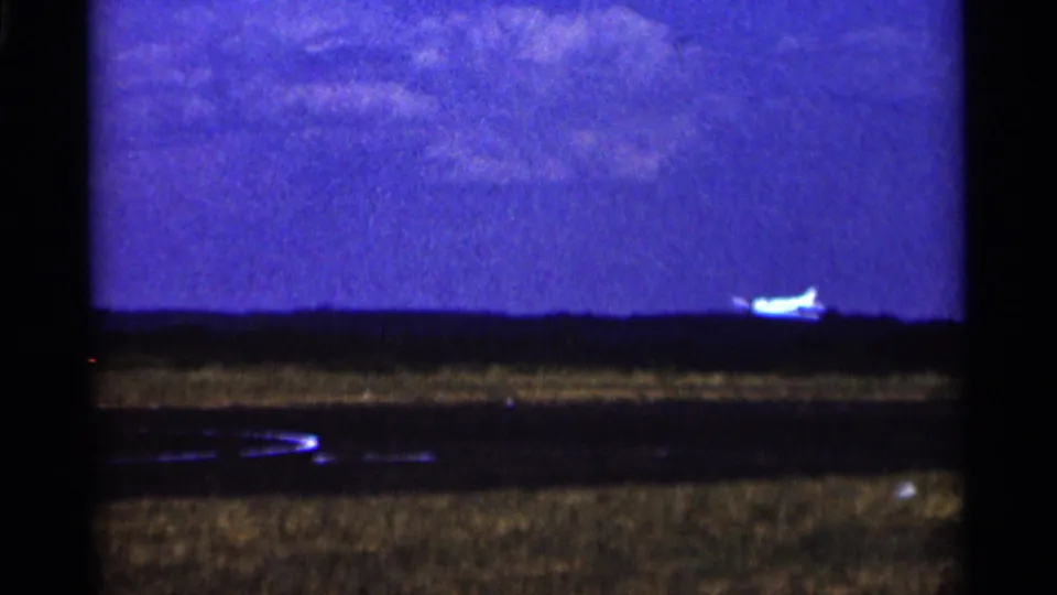 1964 Minnesota 8mm frame showing a small white airplane near the horizon over a dark treeline, with runway and grass below.