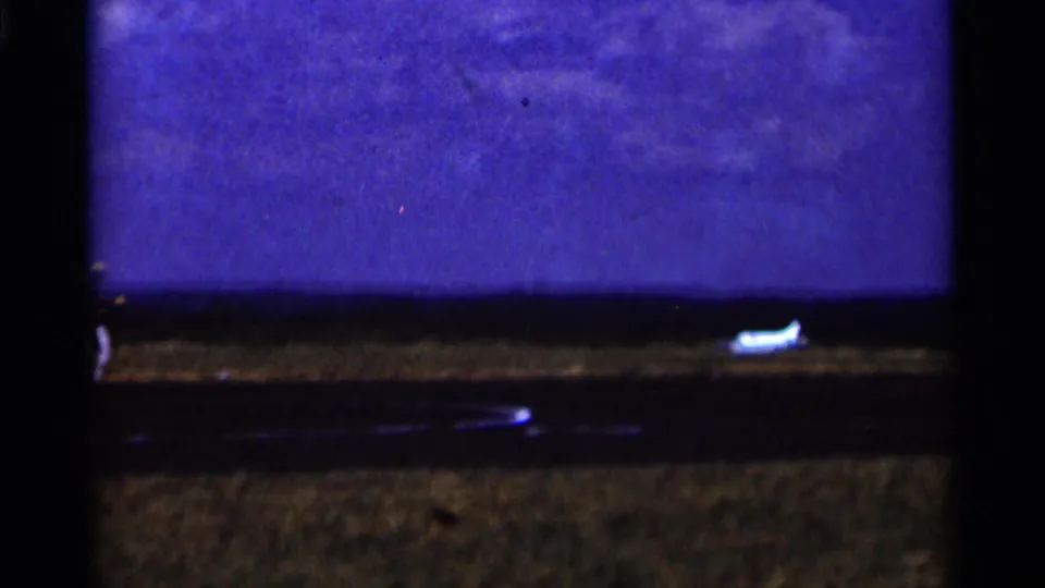 1964 Minnesota 8mm frame showing a small white model airplane near a dark landing strip, with open grass and bright sky.