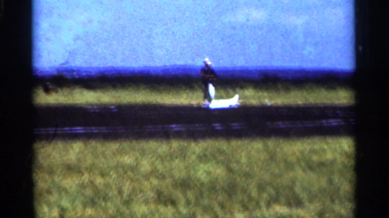 1964 Minnesota home-movie frame with a man near a small white model airplane on a grass-side landing strip.