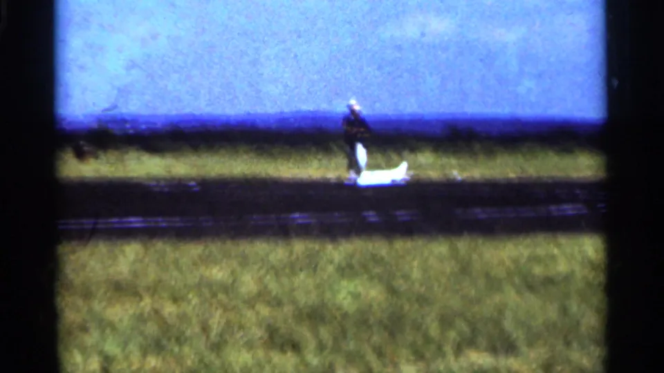 1964 Minnesota home-movie frame with a man near a small white model airplane on a grass-side landing strip.