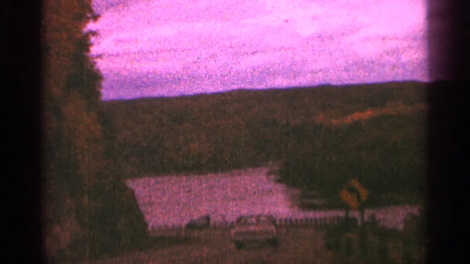 1964 Minnesota roadside view with a lake, tree line, small car, and yellow road sign under a pink sky.
