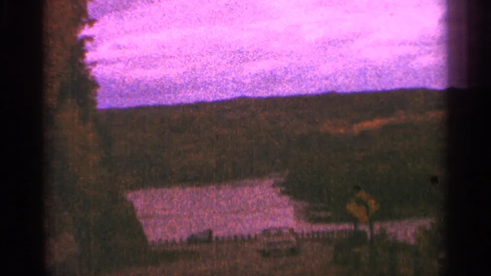 Minnesota home-movie frame showing a small car near a lake, roadside sign, trees, and sunset-colored sky.