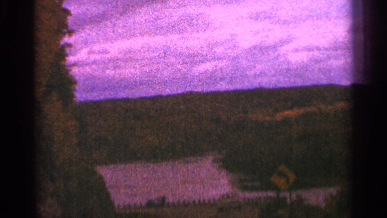 1964 Minnesota 8mm frame with a yellow curve sign, roadside trees, distant water, wooded hills, and strong film grain.