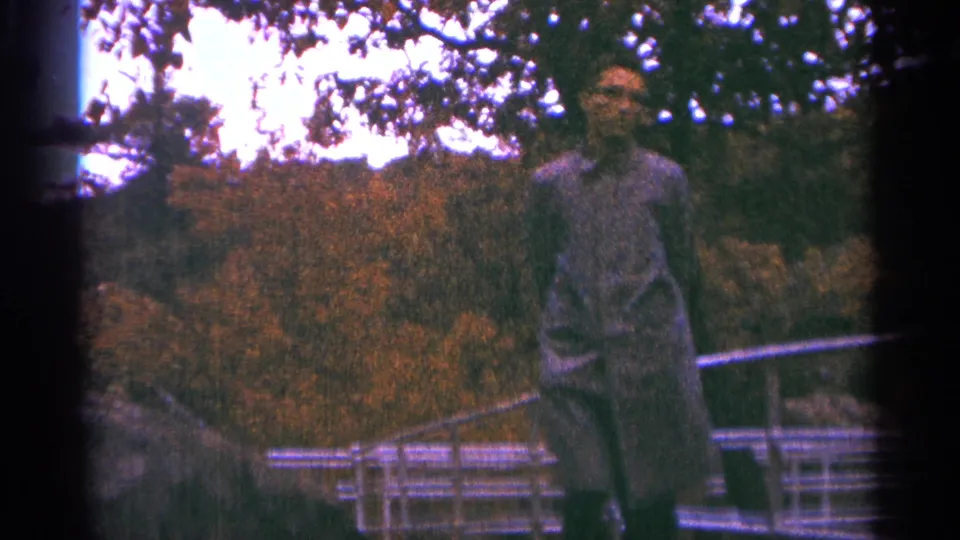 In a 1964 Minnesota park, an adult man walks across a metal bridge amid dense trees and warm-toned foliage.