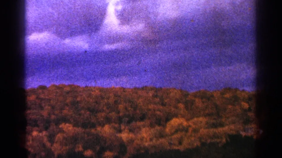 1964 Minnesota home-movie frame with a tree-covered ridge, mottled sky, warm color shift, and dark 8mm frame edge.