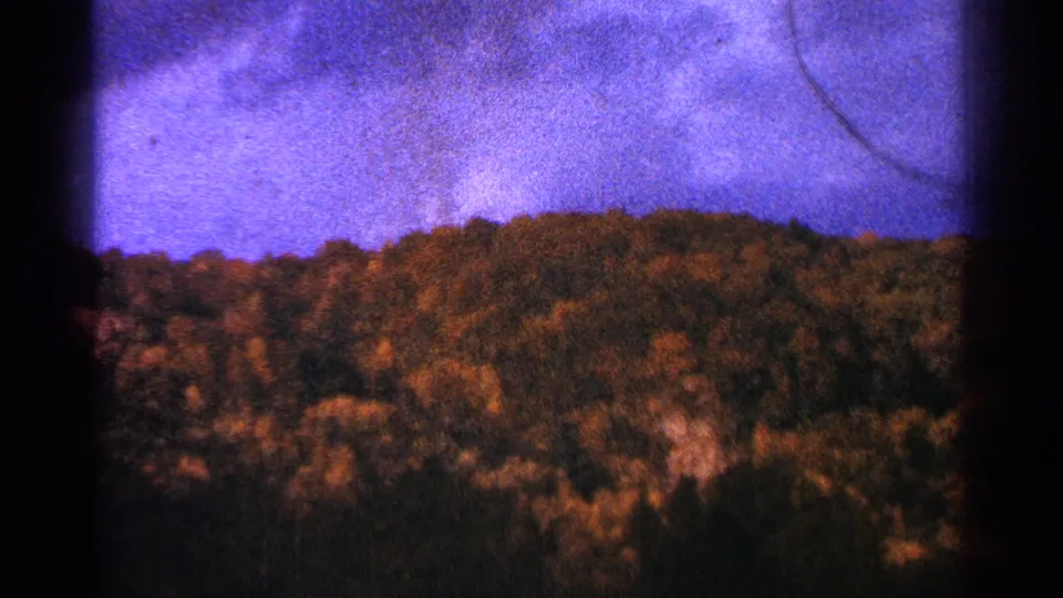 1964 Minnesota 8mm frame of a wooded ridge beneath clouded purple sky, with visible grain, vignetting, and a light scratch.