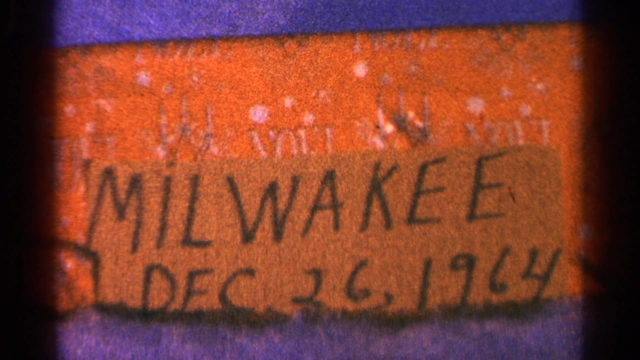 Close 8mm frame of an orange handwritten label reading “MILWAUKEE DEC 26, 1964,” with dark film borders.
