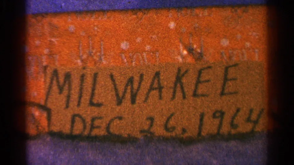 1964 home-movie label frame showing cropped handwriting for Milwaukee and Dec. 26, 1964 on orange cardboard.