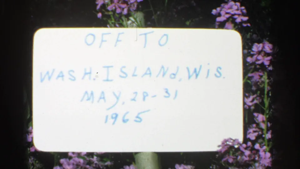 Close 8mm view of the handwritten Washington Island, Wisconsin trip sign, with May dates and purple flowers visible.