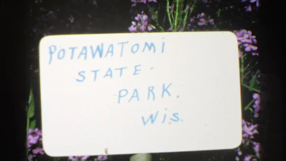 Close-up of a 1964 8mm title card for Potawatomi State Park, Wis., with purple flowers and foliage behind the card.