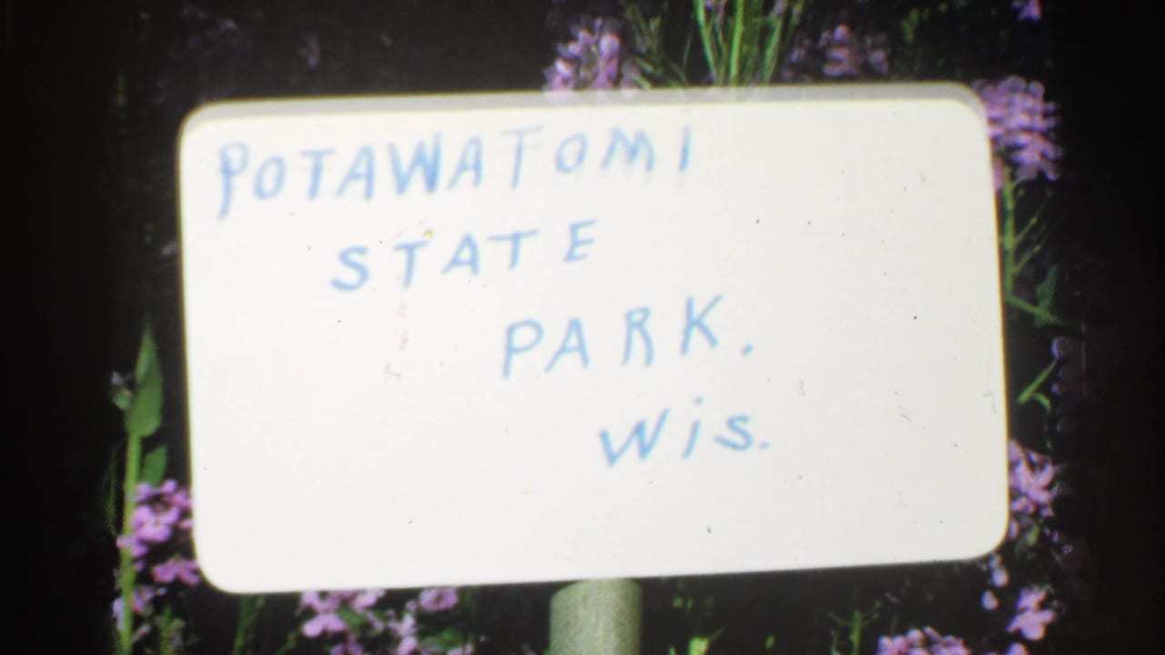 1964 8mm frame showing a handwritten Pottawatomie State Park, Wisconsin title card set against flowers and dark foliage.