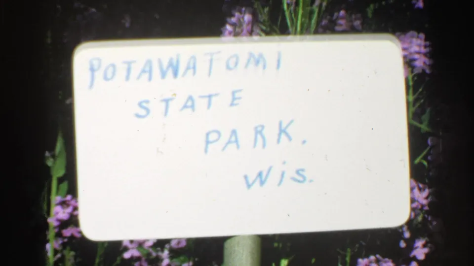 1964 8mm frame showing a handwritten Pottawatomie State Park, Wisconsin title card set against flowers and dark foliage.