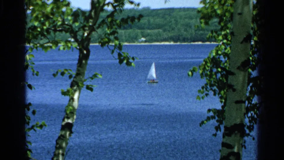 1964 8mm lake view at Pottawatomie State Park, with a small sailboat on blue water and birch trunks framing the scene.