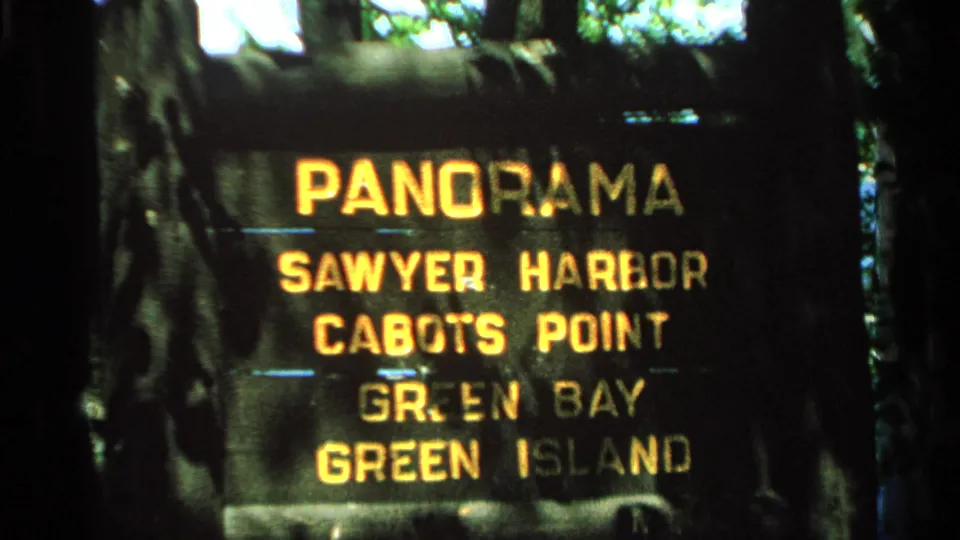 Washington Island home-movie frame from 1964 showing a close-up of a wooden scenic sign with bright yellow destination lettering.