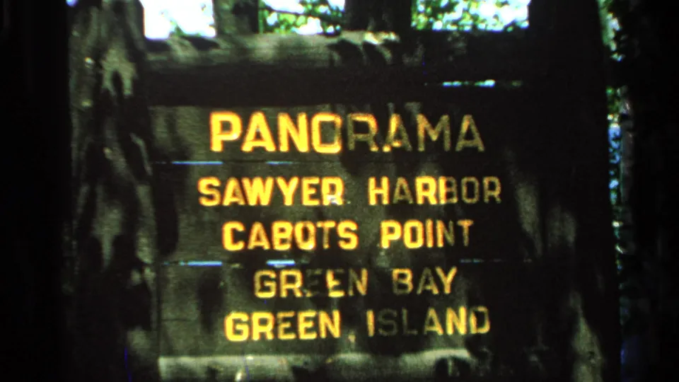 1964 8mm frame of the Washington Island scenic sign, with soft focus, grain, and yellow lettering against dark wood.