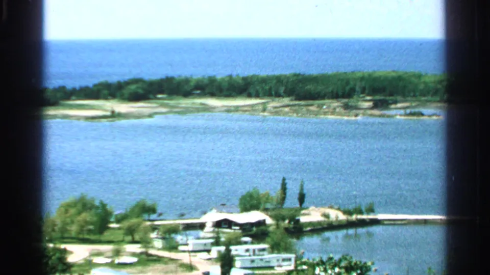 1964 Washington Island 8mm frame showing a sunny lake panorama with blue water, wooded shore, trailers, dock area, and a small waterfront building.