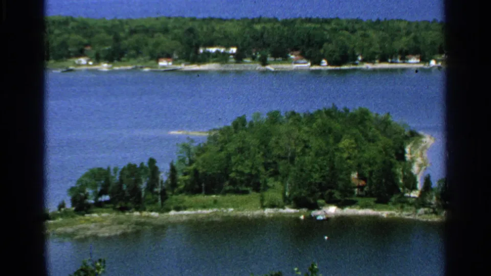 1964 Washington Island 8mm frame showing a tree-covered point, surrounding lake water, and small distant shoreline houses.
