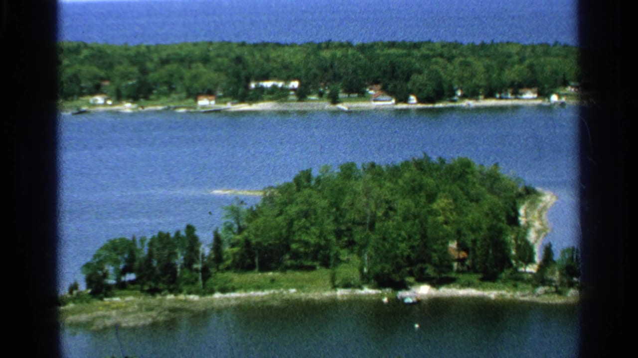 1964 Washington Island, Wisconsin home-movie frame showing a wooded island, lake water, and scattered shoreline homes from above.