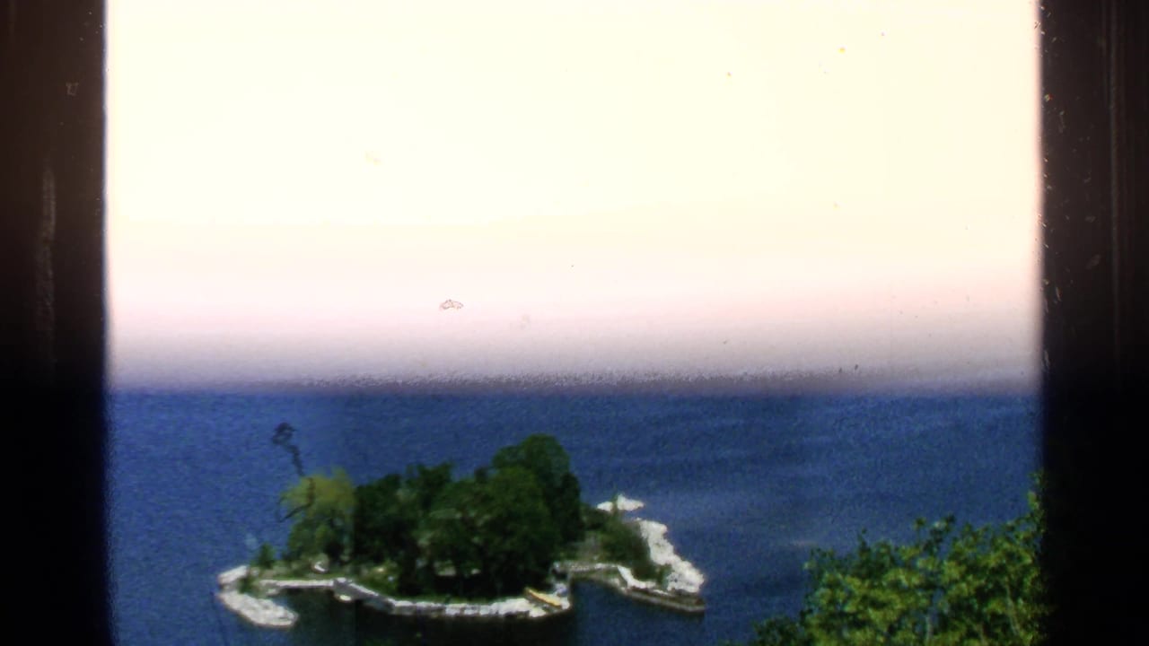 Washington Island-area lake view with a small tree-covered island, curved shoreline, and open blue water in a 1964 8mm frame.