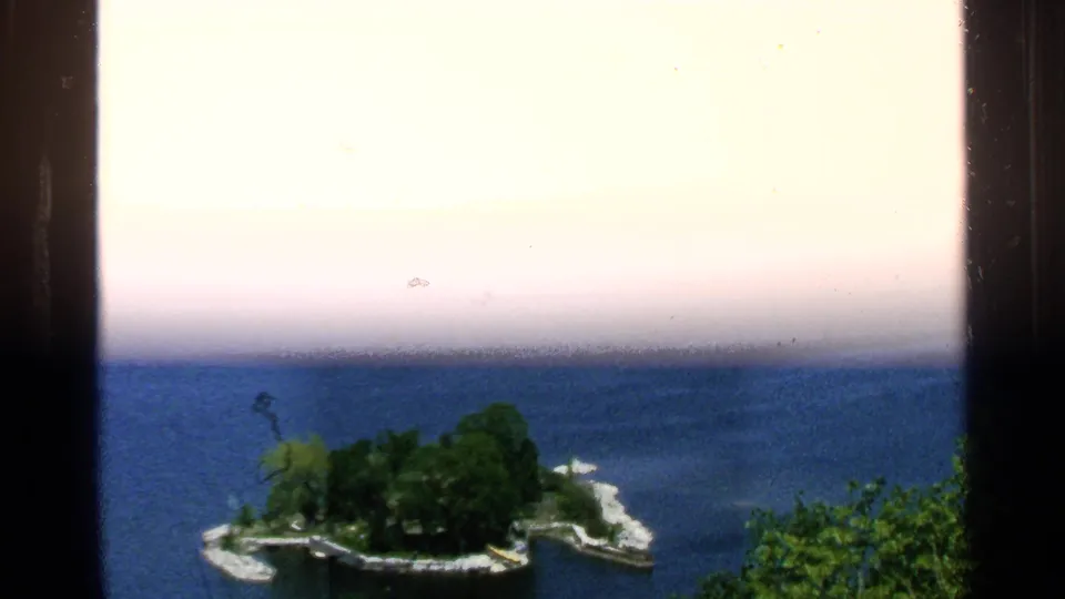 Washington Island-area lake view with a small tree-covered island, curved shoreline, and open blue water in a 1964 8mm frame.
