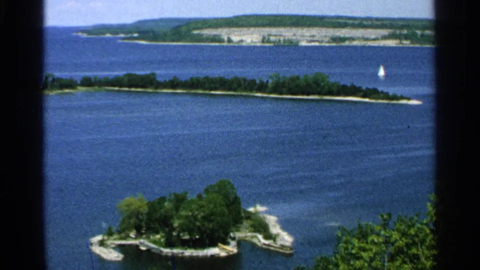 1964 elevated Washington Island lake panorama showing a wooded foreground island, distant shoreline, and a small white sailboat.