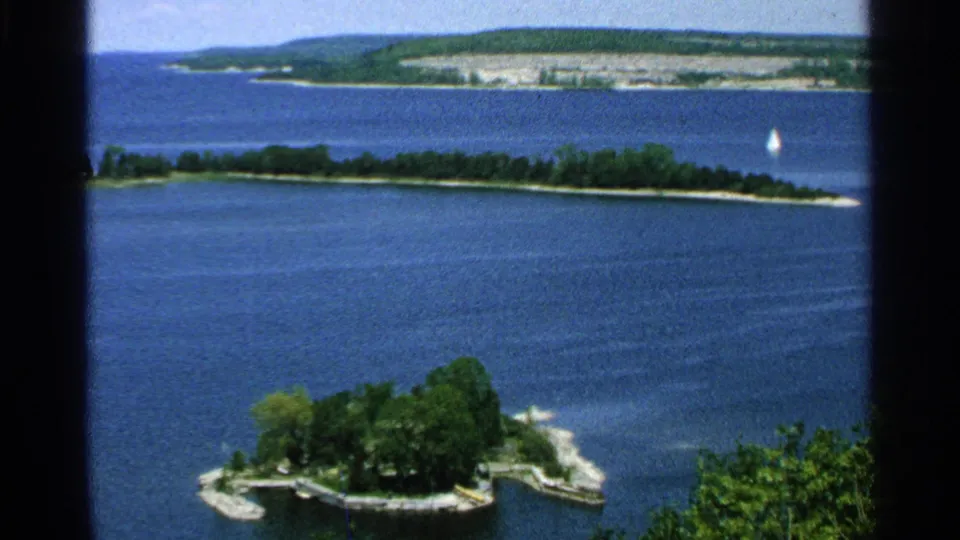 Elevated 1964 Washington Island view with blue water, a wooded island, distant shore, and a small sailboat near the horizon line.