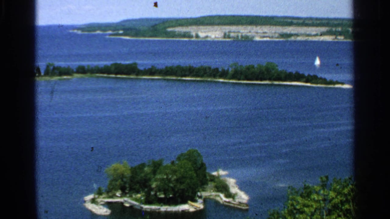 1964 Washington Island 8mm view across blue lake water toward a small wooded islet, distant shore, and tiny sailboat.