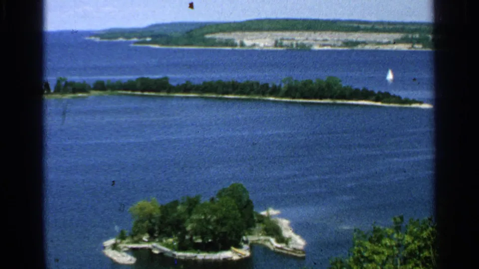 1964 Washington Island 8mm view across blue lake water toward a small wooded islet, distant shore, and tiny sailboat.