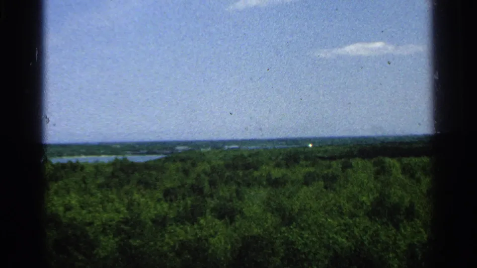 1964 8mm view from Washington Island over a wooded landscape, distant water, and a bright sky with a small cloud.
