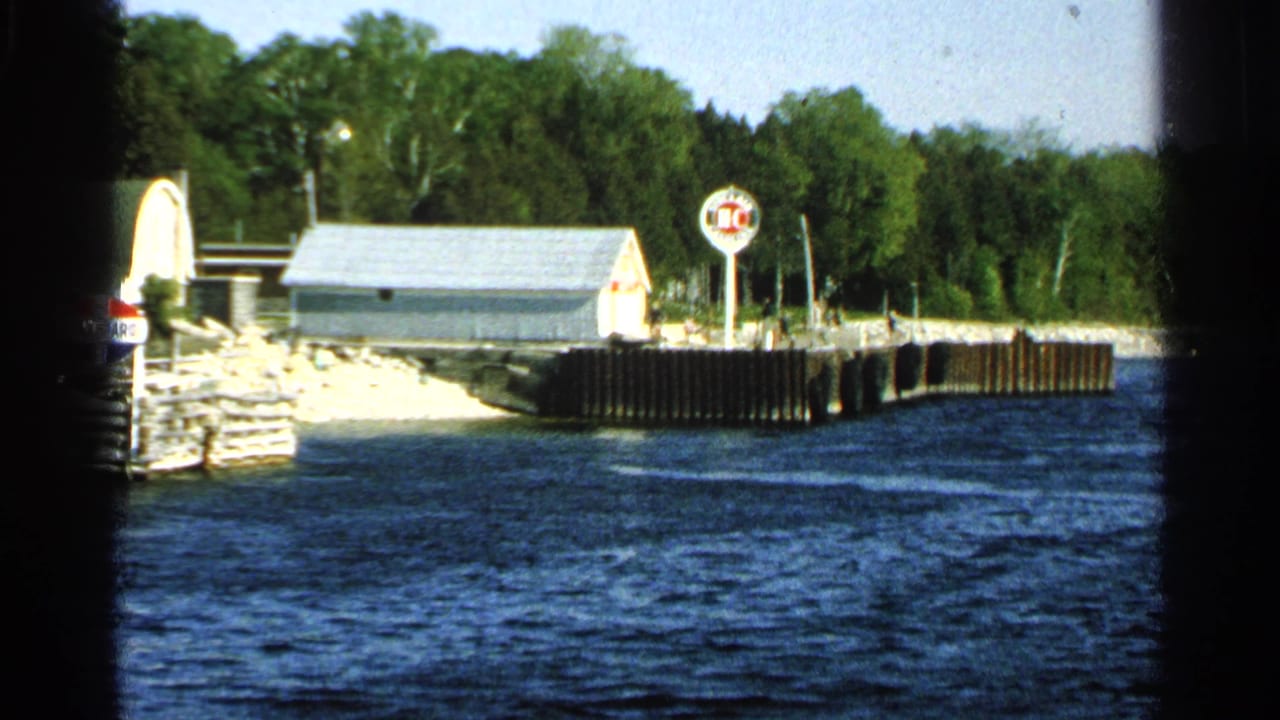 Waterfront dock with small buildings, signs, blue water, and trees behind the shore.